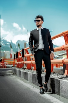 A man in a stylish black suit and sunglasses stands confidently beside an orange safety barrier on a road. The background features a mountainous landscape under a clear blue sky.
