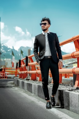 A man in a stylish black suit and sunglasses stands confidently beside an orange safety barrier on a road. The background features a mountainous landscape under a clear blue sky.
