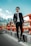A man in a stylish black suit and sunglasses stands confidently beside an orange safety barrier on a road. The background features a mountainous landscape under a clear blue sky.