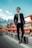 A man in a stylish black suit and sunglasses stands confidently beside an orange safety barrier on a road. The background features a mountainous landscape under a clear blue sky.
