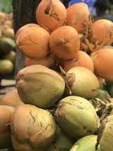 A pile of coconuts with various shades of green and orange, featuring rough textures and some natural blemishes. The coconuts are clustered closely together, suggesting a tropical setting.