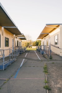 A row of portable classroom buildings with ramps and railings, set against a background of trees and open sky. The path between the buildings is paved with cracks, and there are small patches of grass and weeds. The setting appears to be outdoors in a school or educational campus.