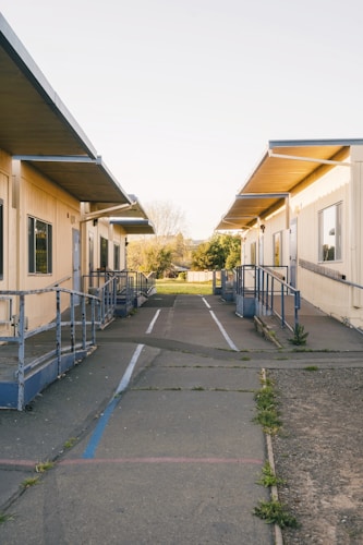 A row of portable classroom buildings with ramps and railings, set against a background of trees and open sky. The path between the buildings is paved with cracks, and there are small patches of grass and weeds. The setting appears to be outdoors in a school or educational campus.