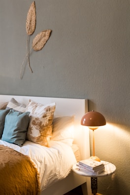 A cozy bedroom corner with a neatly made bed adorned with an array of pillows in various patterns and textures. A soft, warm blanket is spread across the bed. A small, round side table beside the bed holds a stack of books underneath a retro-style lamp casting a warm glow. Above the headboard, two textured leaf decorations are mounted on the wall, adding a natural element to the room’s decor.