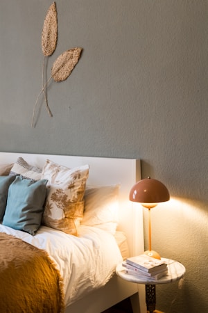 A cozy bedroom corner with a neatly made bed adorned with an array of pillows in various patterns and textures. A soft, warm blanket is spread across the bed. A small, round side table beside the bed holds a stack of books underneath a retro-style lamp casting a warm glow. Above the headboard, two textured leaf decorations are mounted on the wall, adding a natural element to the room&rsquo;s decor.