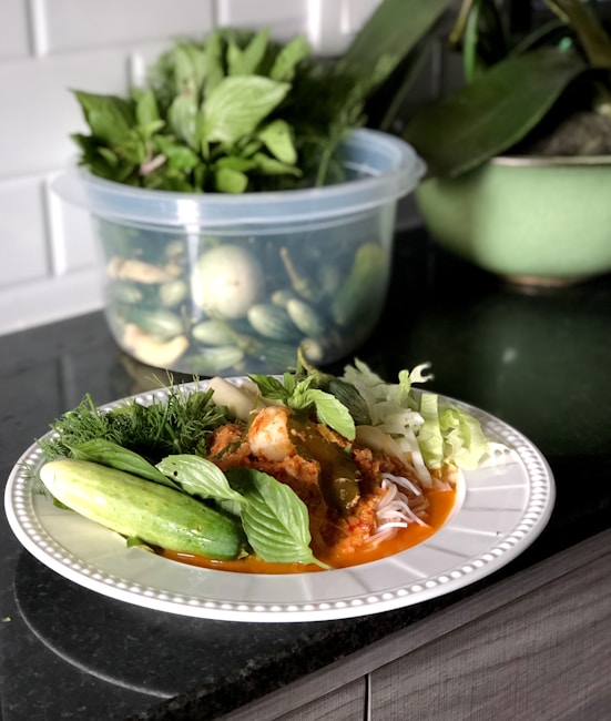 A plate of food sits on a countertop, featuring a vibrant red sauce with noodles and garnished with fresh herbs and vegetables. In the background, a plastic container holds an assortment of green vegetables and herbs, contributing to the fresh, garden-like atmosphere.