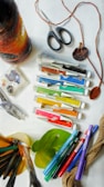 A collection of art supplies is scattered on a surface, including tubes of paint, markers, a pair of scissors, yarn, leaves, and a paintbrush. There is a wrapped bottle with yarn in gradient colors from black to orange. The scene suggests a creative workspace with various tools for crafting or painting.