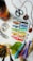 A collection of art supplies is scattered on a surface, including tubes of paint, markers, a pair of scissors, yarn, leaves, and a paintbrush. There is a wrapped bottle with yarn in gradient colors from black to orange. The scene suggests a creative workspace with various tools for crafting or painting.