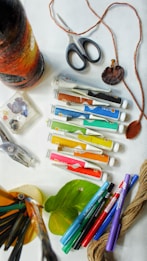 A collection of art supplies is scattered on a surface, including tubes of paint, markers, a pair of scissors, yarn, leaves, and a paintbrush. There is a wrapped bottle with yarn in gradient colors from black to orange. The scene suggests a creative workspace with various tools for crafting or painting.