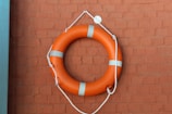 An orange life preserver with white stripes is hanging on a textured brick wall. The wall is painted in a warm brownish-red color and the life preserver is secured with white ropes.
