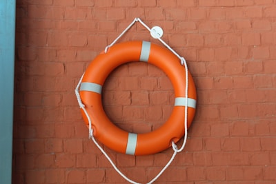 An orange life preserver with white stripes is hanging on a textured brick wall. The wall is painted in a warm brownish-red color and the life preserver is secured with white ropes.