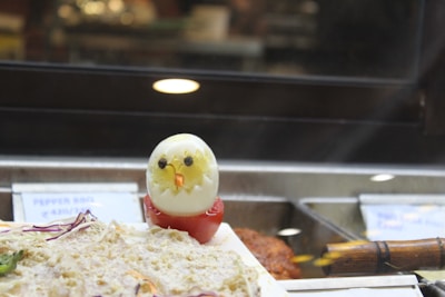 A creatively designed egg resembles a chicken, with black olives for eyes and a piece of carrot for the beak, placed atop a halved tomato. The setup is in a deli or buffet setting with various food items displayed, including a price tag visible in the background.