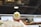 A creatively designed egg resembles a chicken, with black olives for eyes and a piece of carrot for the beak, placed atop a halved tomato. The setup is in a deli or buffet setting with various food items displayed, including a price tag visible in the background.