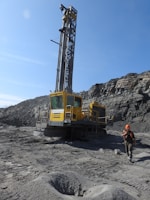 A large industrial drilling machine is positioned in a rocky, barren landscape. The machine is predominantly yellow with visible branding and is equipped for deep drilling. A person wearing a hard hat and protective gear is walking nearby, highlighting an industrial or mining environment. The sky above is clear and blue.