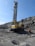 A large industrial drilling machine is positioned in a rocky, barren landscape. The machine is predominantly yellow with visible branding and is equipped for deep drilling. A person wearing a hard hat and protective gear is walking nearby, highlighting an industrial or mining environment. The sky above is clear and blue.