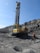 A large industrial drilling machine is positioned in a rocky, barren landscape. The machine is predominantly yellow with visible branding and is equipped for deep drilling. A person wearing a hard hat and protective gear is walking nearby, highlighting an industrial or mining environment. The sky above is clear and blue.