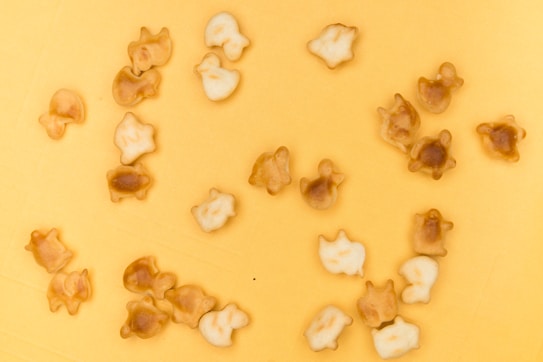 A collection of small, biscuit-like snacks shaped like various animals, scattered across a yellow surface. The snacks have a light brown and golden color, some darker than others, suggesting varying levels of baking.