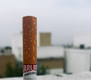A close-up view of a cigarette held vertically with a blurred urban background of buildings and a cloudy sky. The cigarette has a brown and white body with visible text near the filter.