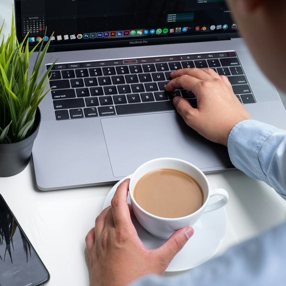 person holding white ceramic mug on macbook pro