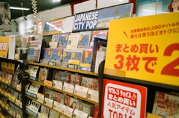 A music store shelf is filled with various CDs, focusing on a section labeled 'Japanese City Pop.' The area is well-organized with different discs prominently displaying labels such as 'Disc 1,' 'Disc 2,' and 'Disc 3.' Promotional signage in Japanese is visible, offering discounts. Headphones and numerous CD covers are present, creating a vibrant and busy atmosphere.
