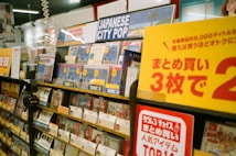 A music store shelf is filled with various CDs, focusing on a section labeled 'Japanese City Pop.' The area is well-organized with different discs prominently displaying labels such as 'Disc 1,' 'Disc 2,' and 'Disc 3.' Promotional signage in Japanese is visible, offering discounts. Headphones and numerous CD covers are present, creating a vibrant and busy atmosphere.
