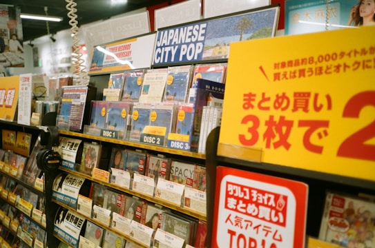 A music store shelf is filled with various CDs, focusing on a section labeled 'Japanese City Pop.' The area is well-organized with different discs prominently displaying labels such as 'Disc 1,' 'Disc 2,' and 'Disc 3.' Promotional signage in Japanese is visible, offering discounts. Headphones and numerous CD covers are present, creating a vibrant and busy atmosphere.