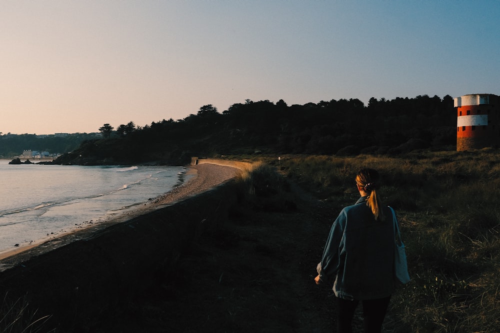 woman in blue jacket standing on brown dirt road near body of water during daytime