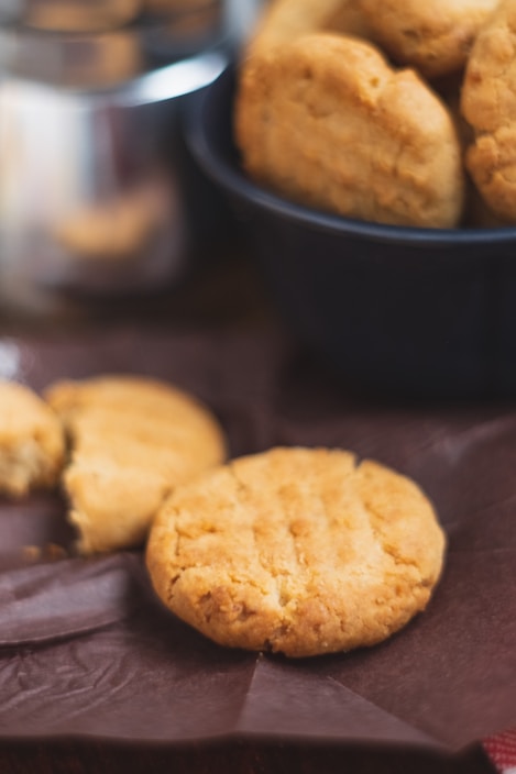 Several round cookies are placed in and around a dark blue bowl. The cookies have a golden brown color and a crumbly texture. One cookie is prominently in the foreground on a piece of brown parchment paper, while others are in the background.