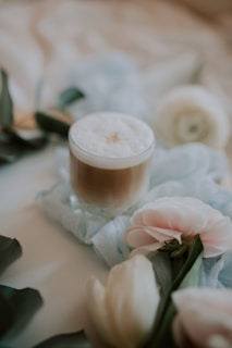 A creamy, frothy beverage sits in a glass on a soft fabric surface surrounded by delicate flowers. The atmosphere is serene and calming.