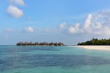 A serene beach scene with clear blue skies and scattered white clouds. Overwater bungalows sit on stilts, extending from a sandy white beach lined with lush green palm trees. The turquoise ocean is calm and inviting.