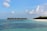 A serene beach scene with clear blue skies and scattered white clouds. Overwater bungalows sit on stilts, extending from a sandy white beach lined with lush green palm trees. The turquoise ocean is calm and inviting.