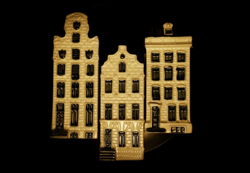 The image features three ceramic models of traditional, narrow, European-style buildings with decorative facades and multiple windows arranged in a row against a dark background. The buildings have a textured surface and are predominantly beige with black details.