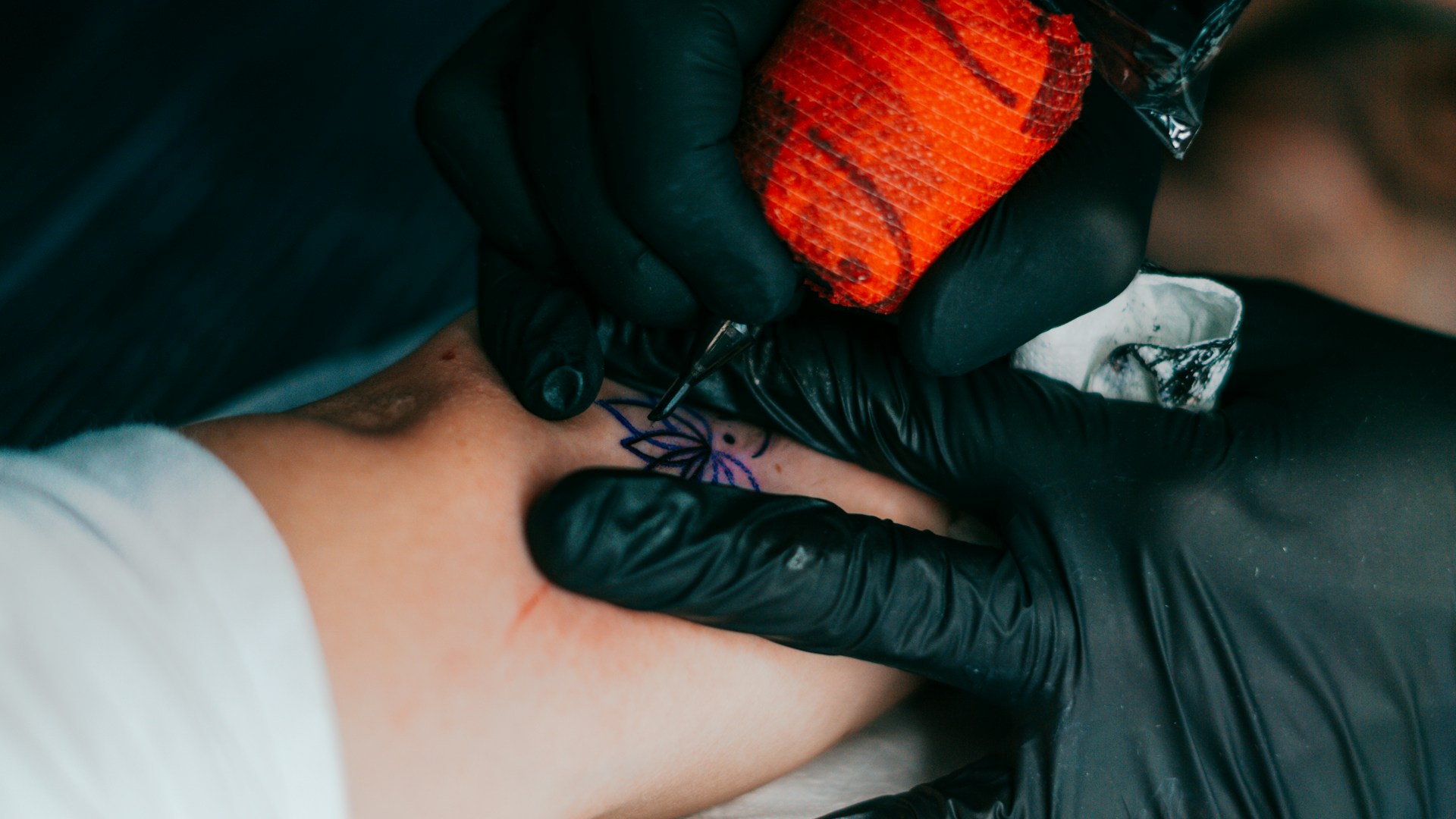 Close-up of a tattoo artist's hand carefully drawing a custom design with a tattoo machine, surrounded by red and black ink bottles.