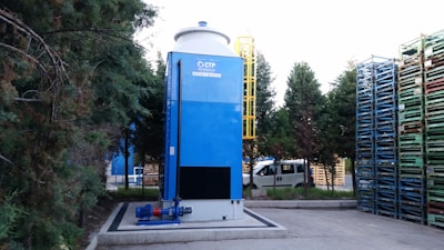 A large blue industrial tower stands next to a stack of multicolored metal pallets. Trees surround the area, providing a natural backdrop. A white van is parked nearby, indicating an industrial or warehouse setting. There is a yellow ladder attached to the tower for maintenance access.