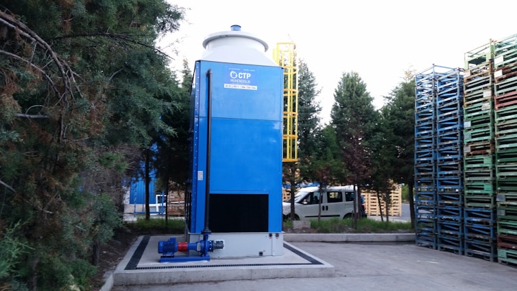 A large blue industrial tower stands next to a stack of multicolored metal pallets. Trees surround the area, providing a natural backdrop. A white van is parked nearby, indicating an industrial or warehouse setting. There is a yellow ladder attached to the tower for maintenance access.