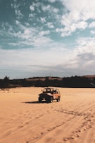 A rugged off-road vehicle is driving across expansive sandy dunes under a partly cloudy sky. The landscape is open and arid, bordered by silhouetted trees in the background.
