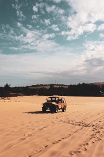 A rugged off-road vehicle is driving across expansive sandy dunes under a partly cloudy sky. The landscape is open and arid, bordered by silhouetted trees in the background.