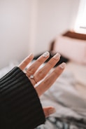 A close-up of a hand with a black sweater sleeve partially covering the wrist. The fingernails are manicured, featuring light colors and a decorative accent with gemstones on at least one nail.