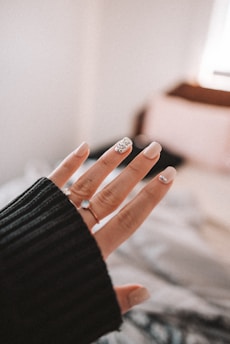 A close-up of a hand with a black sweater sleeve partially covering the wrist. The fingernails are manicured, featuring light colors and a decorative accent with gemstones on at least one nail.