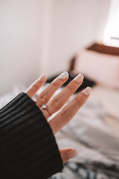 A close-up of a hand with a black sweater sleeve partially covering the wrist. The fingernails are manicured, featuring light colors and a decorative accent with gemstones on at least one nail.