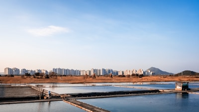 A scenic landscape featuring a series of rectangular water bodies, possibly salt pans, in the foreground. Beyond them, there's a flat expanse of dry grassland, and a skyline with modern high-rise buildings stretching across the horizon. A mountain is visible in the background under a clear blue sky, adding depth to the composition.