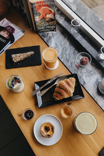 An assortment of breakfast items is laid out on a wooden table near a window. A book titled 'The Sea Change' is propped up next to a stack of magazines. There is a croissant on a black rectangular plate with a knife and fork, a slice of cake on a slate board, a small glass of yogurt with berries, a jam jar, a custard tart, a glass of coffee, and a smoothie. The scene is casual and inviting, suggesting a relaxed, cozy breakfast setting.