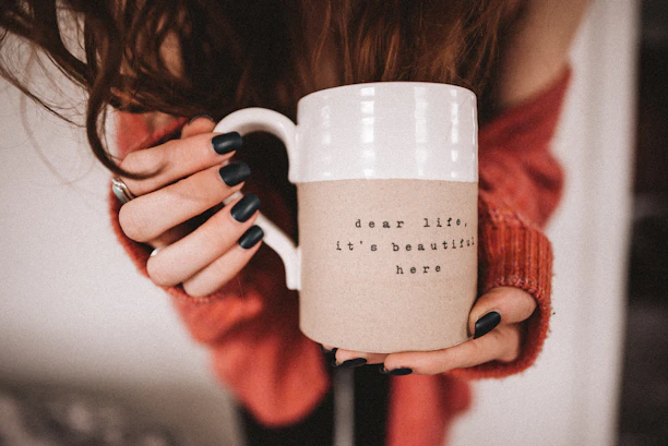 Close-up of a white mug featuring a witty African proverb, held by a smiling hand with earthy-toned nail polish.