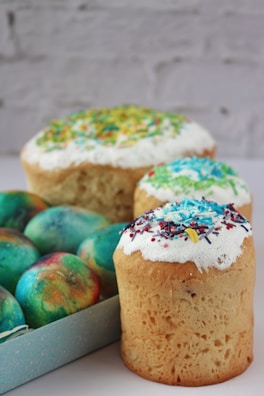 Three decorated Easter cakes with colorful sprinkles on top are placed next to a box of vibrantly colored dyed eggs. The cakes have a fluffy texture and are topped with white icing covered in sprinkles. The eggs display a variety of teal, blue, and orange hues, giving a festive appearance.