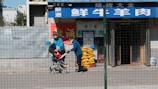 A person wearing a blue jacket and a face mask pushes a child in a stroller along a sidewalk. They pass in front of a storefront with blue and white signage in a foreign language and a red banner with yellow text. On the side of the store, there are stacked yellow items, possibly bags. A wire fence runs in front of the scene, partially obscuring the view.
