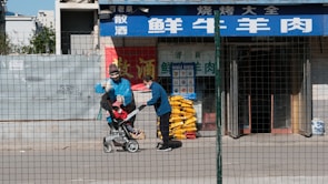 A person wearing a blue jacket and a face mask pushes a child in a stroller along a sidewalk. They pass in front of a storefront with blue and white signage in a foreign language and a red banner with yellow text. On the side of the store, there are stacked yellow items, possibly bags. A wire fence runs in front of the scene, partially obscuring the view.