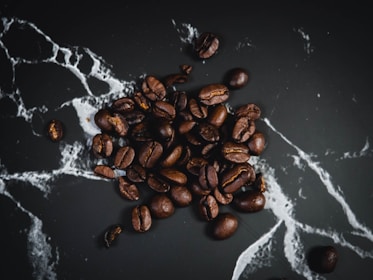 A collection of roasted coffee beans scattered on a dark, textured surface with white, marble-like veins running through it.