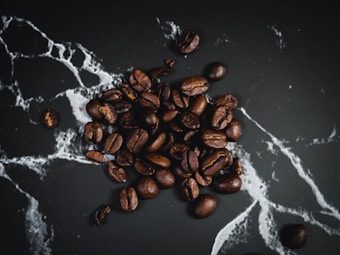 A collection of roasted coffee beans scattered on a dark, textured surface with white, marble-like veins running through it.