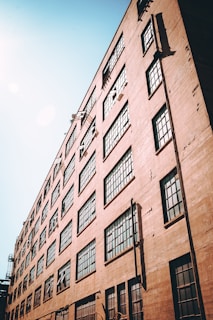 A tall, industrial-style building with a series of large, rectangular windows. The facade is made of a beige or light brown material, and the sunlight casts sharp shadows across the surface. The image is taken from an upward angle, emphasizing the height of the building against a clear blue sky.