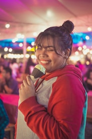 A person with a joyful expression holds an ice cream cone, wearing a red and gray hoodie. The background is a lively outdoor setting with colorful lights and blurred people.